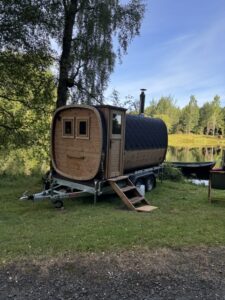 Mobil Hordó Kabin Szauna 87 A TimberIN Nordic Square mobil szauna oldalnézete utánfutón, kéménnyel egy tó mellett